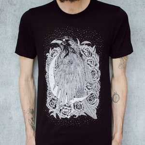A unisex black t-shirt with a white hand screen printed design. The detailed hand drawn natural illustration is of a raven against a large crescent moon and peony flowers over a starry sky.