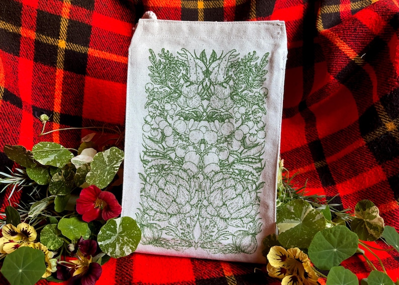 A natural colored canvas lunchbag featuring hand drawn art of various vegetables including artichokes, radishes, tomatoes, peas, chanterelle mushrooms, and garlic. The design is screen printed in warm green pink on the front of the bag.