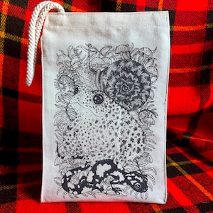 An off white natural canvas lunch bag with a rope handle and a deep charcoal grey hand screen printed design. The hand drawn natural illustration on the bag features a toad, a snail, a spotted salamander, ferns, and small mushrooms.