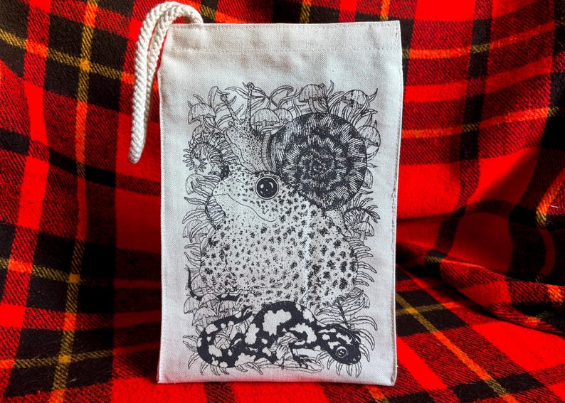 An off white natural canvas lunch bag with a rope handle and a deep charcoal grey hand screen printed design. The hand drawn natural illustration on the bag features a toad, a snail, a spotted salamander, ferns, and small mushrooms.