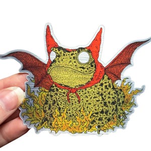 A die cut brushed silver metallic sticker of a full color toad in a devil costume with wings and flames.