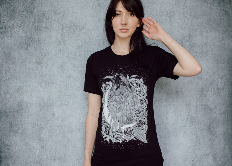 A unisex black t-shirt with a white hand screen printed design. The detailed hand drawn natural illustration is of a raven against a large crescent moon and peony flowers over a starry sky.