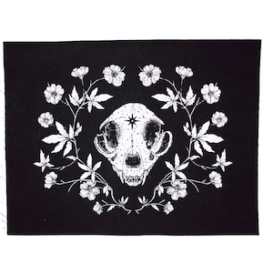 May include: Black fabric patch featuring a white illustration of a cat skull encircled by a wreath of white flowers and leaves.