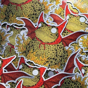 A die cut brushed silver metallic sticker of a full color toad in a devil costume with wings and flames.