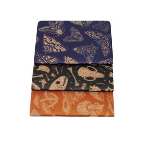 A set of three kraft cardstock pocket notebooks with natural illustration patters on the outside. One is print in blue ink with moths, one is black ink with skulls, and the third is red ink with various mushrooms and fungi.