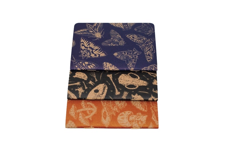 A set of three kraft cardstock pocket notebooks with natural illustration patters on the outside. One is print in blue ink with moths, one is black ink with skulls, and the third is red ink with various mushrooms and fungi.