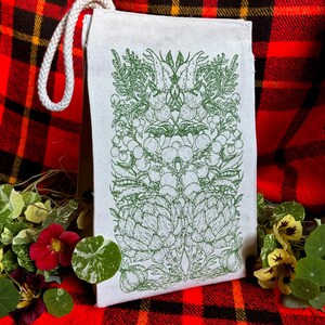 A natural colored canvas lunchbag featuring hand drawn art of various vegetables including artichokes, radishes, tomatoes, peas, chanterelle mushrooms, and garlic. The design is screen printed in warm green pink on the front of the bag.