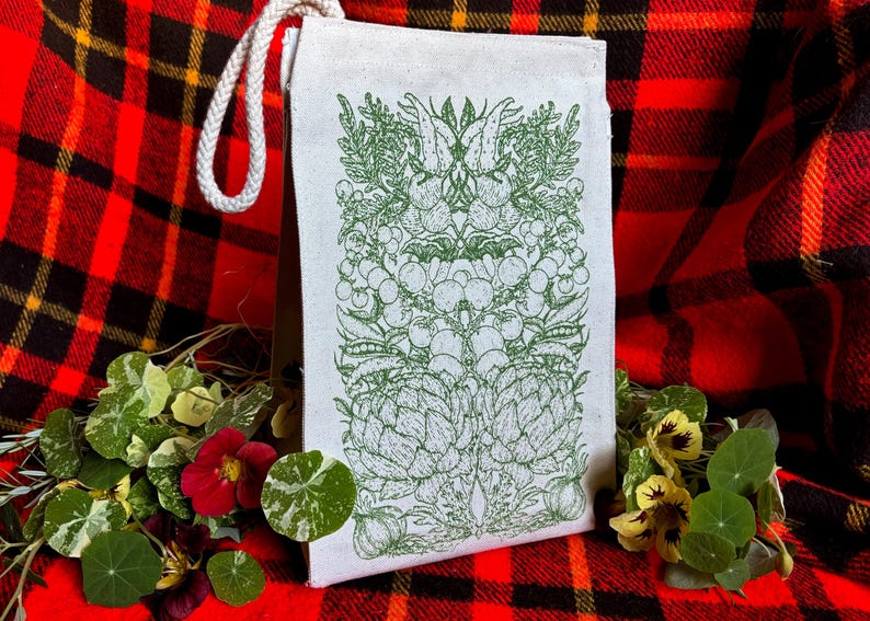 A natural colored canvas lunchbag featuring hand drawn art of various vegetables including artichokes, radishes, tomatoes, peas, chanterelle mushrooms, and garlic. The design is screen printed in warm green pink on the front of the bag.
