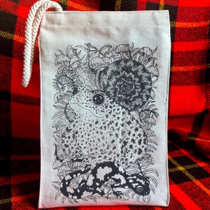 An off white natural canvas lunch bag with a rope handle and a deep charcoal grey hand screen printed design. The hand drawn natural illustration on the bag features a toad, a snail, a spotted salamander, ferns, and small mushrooms.