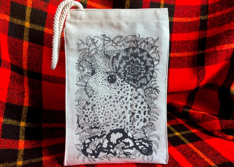 An off white natural canvas lunch bag with a rope handle and a deep charcoal grey hand screen printed design. The hand drawn natural illustration on the bag features a toad, a snail, a spotted salamander, ferns, and small mushrooms.