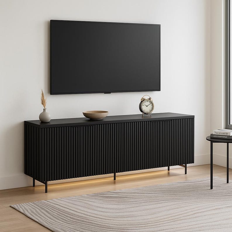 May include: A modern black entertainment center with a ribbed front design, supporting a large flat-screen TV. The cabinet has a minimalist aesthetic, with a small vase, bowl, and clock on top. The cabinet is 72 inches long.