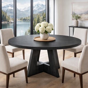 May include: A dark gray round dining table with a pedestal base and four beige upholstered chairs. A white vase with white flowers sits on a wooden tray in the center of the table. The background shows a scenic view of a lake and mountains.