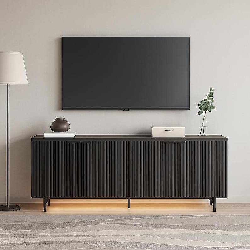 May include: A dark brown modern TV stand with vertical grooved detailing. The stand has a rectangular shape with four legs and a light strip underneath. A flat-screen TV is mounted above the stand, with decorative items on top.