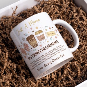 May include: White ceramic mug with the text "Just A Mama" in gold script. The mug features illustrations of coffee cups, a bow, and the text "#STRINGCHEESEMAMA". The mug is inside a white box with brown paper filler.