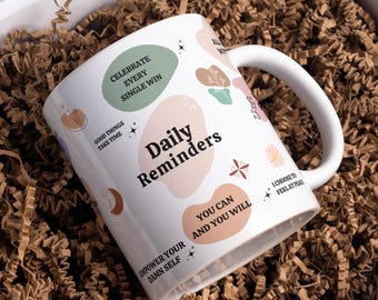May include: White ceramic mug with the words "Daily Reminders" and inspirational phrases like "Celebrate Every Single Win" and "Empower Your Damn Self." The mug has a white handle and is surrounded by brown packing material.