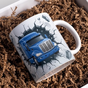 Puede incluir: Taza de cerámica blanca con un diseño de camión azul rompiendo una pared agrietada. La taza tiene un asa blanca y está rodeada de material de embalaje marrón.