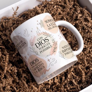 May include: A white ceramic mug featuring various Spanish phrases and illustrations including candles, doves, and floral accents. The text includes "CON DIOS TODO ES POSIBLE," "SOMOS LA LUZ DEL MUNDO," "DIOS ES MI FORTALEZA," "NADA ES IMPOSIBLE PARA DIOS," and "DIOS TIENE MEJORES CAFECITO Y A JESÚS."