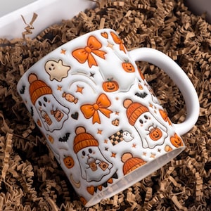 Puede incluir: Taza de cerámica blanca con un diseño temático de Halloween. El diseño incluye fantasmas de dibujos animados con gorros naranjas, calabazas, lazos y murciélagos. La taza tiene un asa blanca y está en una caja con relleno de papel marrón.