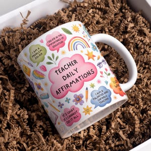 May include: White ceramic mug with the text "Teacher Daily Affirmations" in pink. The mug is decorated with colorful illustrations and affirmations, including "I am a problem solver" and "I believe in the importance of education."