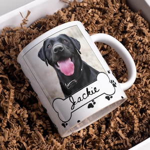 Könnte beinhalten: Weißer Keramikbecher mit einem Foto eines schwarzen Labrador Retrievers. Der Becher hat ein knochenförmiges Design mit dem Namen "Jackie" und Pfotenabdrücken. Der Becher befindet sich in einer weißen Box mit braunem Papierfüllmaterial.