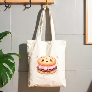 May include: Cream-colored tote bag featuring a cartoon illustration of a smiling cookie sandwich with pink filling and chocolate layers. The bag has long handles and is hanging against a gray wall. Small pink hearts surround the cookie sandwich.