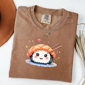 May include: Brown t-shirt with a sushi graphic. The sushi illustration features a smiling face, shrimp, and chopsticks. The shirt has a crew neck and the brand name "COMFY COLORS" is visible on the tag.