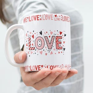May include: White ceramic mug with the word "LOVE" in pink and red, decorated with hearts and the word "LOVE" around the rim. The mug is held by a person.