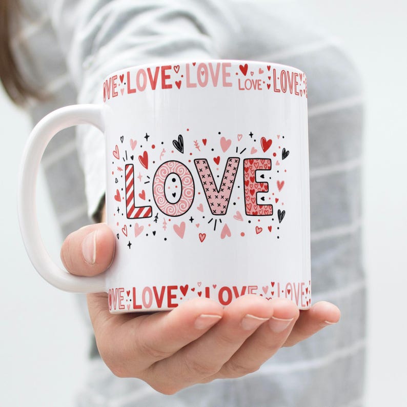 May include: White ceramic mug with the word "LOVE" in pink and red, decorated with hearts and the word "LOVE" around the rim. The mug is held by a person.