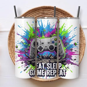 May include: Three white insulated tumblers with black straws, each featuring a colourful design. The design includes a grey video game controller, a splash of blue, green, and purple paint, and the text "EAT SLEEP GAME REPEAT."