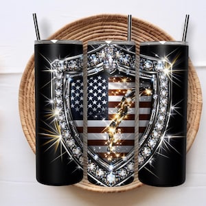May include: Black insulated tumbler with a shield design featuring the American flag and sparkling embellishments. The tumbler has a black straw and is designed to keep beverages cold or hot. The design is patriotic and decorative.