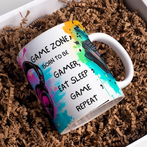 May include: White ceramic mug with the text "GAME ZONE, BORN TO BE GAMER, EAT SLEEP GAME REPEAT" and colorful paint splatter design. The mug features a headset and a keyboard graphic. The mug is inside a white box with brown packing material.