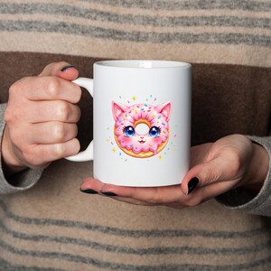 May include: White ceramic mug featuring a colorful illustration of a pink donut designed as a cat. The donut cat has blue eyes, pink ears, and sprinkles. A fun and whimsical design for coffee or tea.
