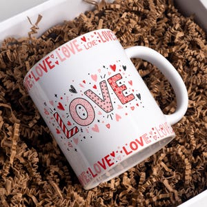 May include: White ceramic mug with the word "LOVE" in pink and red, decorated with hearts and stars. The word "LOVE" is repeated around the rim. The mug is nestled in brown packing material inside a white box.