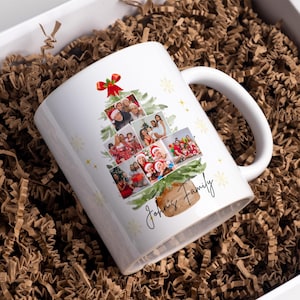 Op de afbeelding: Witte keramische mok met een kerstboomontwerp bestaande uit familiefoto's. De boom is versierd met een rode strik en gouden sterren. De mok is gepersonaliseerd met de tekst "John's Family". De mok zit in een witte geschenkdoos met bruine papieren vulling.
