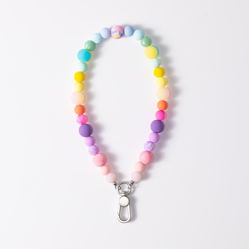 Puede incluir: Un cord&oacute;n circular hecho de cuentas de silicona multicolores en tonos rosa, morado, azul, verde, amarillo y naranja, con una cuenta jaspeada en la parte superior y un mosquet&oacute;n de metal plateado en la parte inferior para su sujeci&oacute;n.