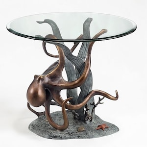 Puede incluir: Una escultura de pulpo de bronce sostiene una mesa redonda de cristal. El pulpo tiene múltiples tentáculos y descansa sobre una base texturizada con una estrella de mar y una concha. El diseño de la mesa incorpora elementos acuáticos.