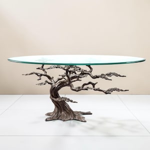 May include: A distinctive coffee table featuring a clear glass top supported by a bronze-coloured metal base shaped like a tree. The base has intricate details mimicking branches and a textured trunk. The table's oval glass top is approximately 122 cm long.