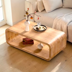 May include: A light-colored wooden coffee table with rounded edges. The table has a lower shelf and is decorated with a white vase, a small tray, and a stack of books. The table is in a living room setting.