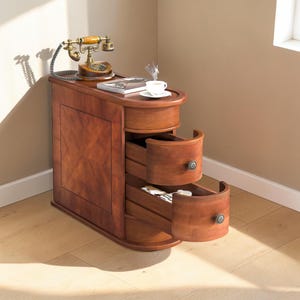 May include: A wooden side table with three drawers, a vintage telephone, and a cup of coffee. The table is a rich brown colour with a patterned front and curved drawer fronts. The drawers have metal knobs.