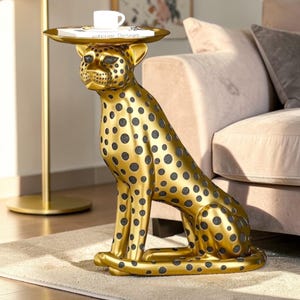 Puede incluir: Una mesa auxiliar dorada con forma de guepardo y manchas negras. El guepardo está sentado, sosteniendo una bandeja redonda sobre su cabeza. Una pequeña taza blanca y un libro están en la bandeja. La mesa está sobre una alfombra clara.