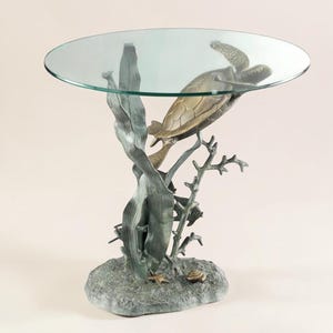 May include: A bronze-colored turtle table with a clear glass top. The table base features a detailed sculpture of a sea turtle, coral, and seaweed. The table is designed to resemble an underwater scene, with a round glass top.