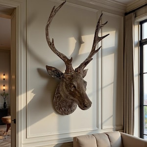 Könnte beinhalten: Eine detaillierte, an der Wand befestigte Hirschkopfskulptur mit großem Geweih. Die Skulptur hat Braun- und Grautöne mit sichtbarer Textur und Detail. Der Kopf ist an einer hellen Wand befestigt und erzeugt einen auffälligen Kontrast.