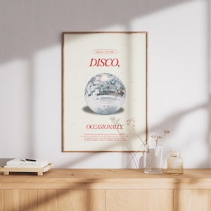 May include: A framed poster with the words "DISCO, OCCASIONALLY" in red text, featuring a mirrored disco ball. The poster is displayed on a wooden surface with a small book and decorative items.