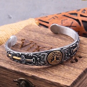 May include: Silver-tone cuff bracelet with intricate Viking-inspired designs. Features a gold-colored tree of life emblem and hammer details. The bracelet is displayed on a wooden box with rope and a decorative wooden piece in the background.
