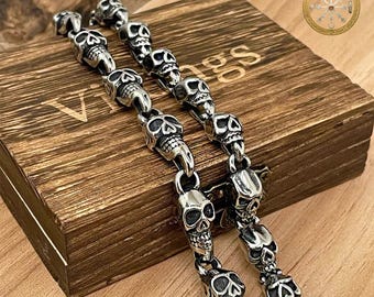 Puede incluir: Dos pulseras plateadas con forma de calavera se muestran sobre una caja de madera. Las pulseras están hechas de pequeños y detallados dijes de calaveras unidos entre sí. La caja tiene la palabra "Vikings" y un logotipo circular con texto visible.
