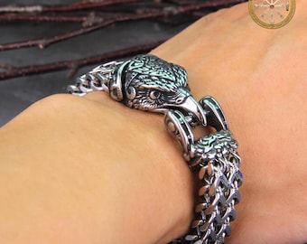 Puede incluir: Una pulsera plateada con un cierre de cabeza de águila. La pulsera tiene un diseño de cadena gruesa. La cabeza del águila está intrincadamente tallada con plumas y un pico afilado. La pulsera se lleva en la muñeca.