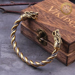 Puede incluir: Un brazalete de estilo vikingo en tonos dorados y plateados con terminales de cabeza de dragón. La banda trenzada es de metal dorado y plateado. El brazalete se exhibe en una caja de madera con las palabras "VIKINGS AMULET" y una rosa de los vientos.
