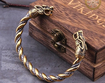 Puede incluir: Un brazalete de estilo vikingo en tonos dorados y plateados con terminales de cabeza de dragón. La banda trenzada es de metal dorado y plateado. El brazalete se exhibe en una caja de madera con las palabras "VIKINGS AMULET" y una rosa de los vientos.