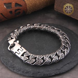 May include: A silver-toned bracelet with an intricate, patterned design. The bracelet features a series of interconnected links, each adorned with swirling, stylized motifs. A sturdy clasp secures the bracelet. The Vikings Amulet logo is visible in the background.