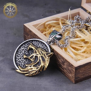 Puede incluir: Un collar con colgante de lobo plateado y dorado. El colgante presenta un diseño detallado de cabeza de lobo sobre un fondo texturizado. El collar se exhibe en una caja de madera con paja. El logotipo The Vikings Amulet está en la esquina superior izquierda.